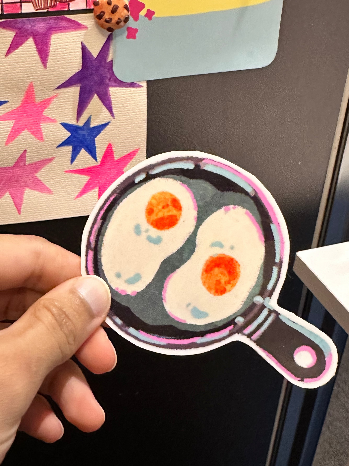 Hand holding a decorative vinyl stickers of fried eggs in a pan against a black surface.