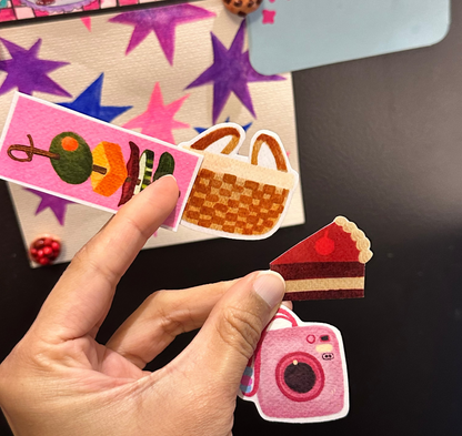 Hand holding 4 small colourful vinyl stickers including a pink camera-shaped sticker, a picnic basket, a snack and a cake slice against a dark surface.
